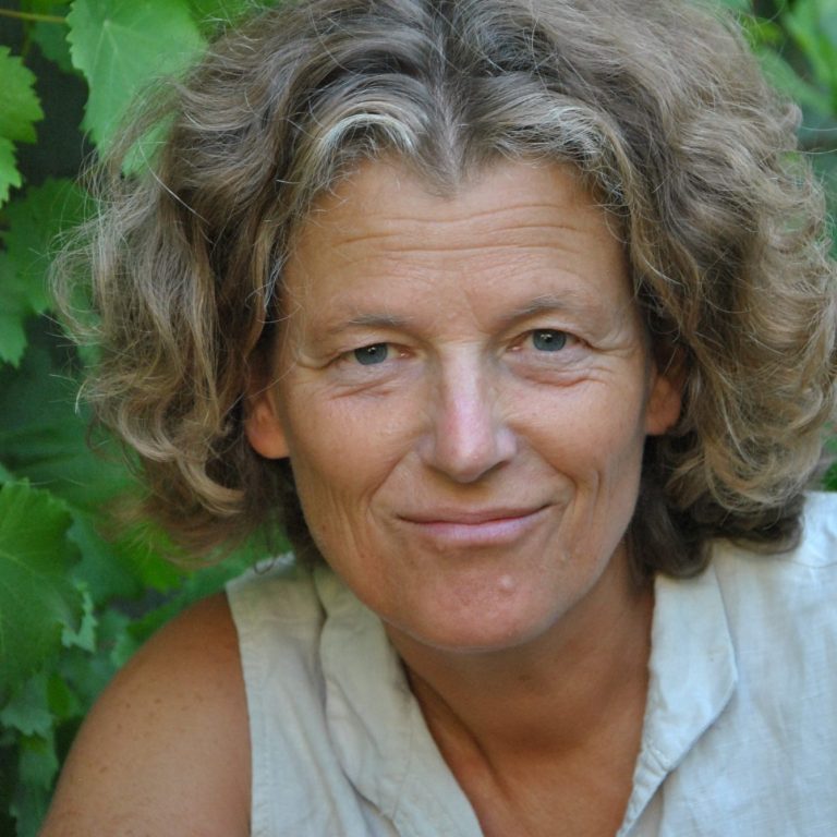 Ursula Riemhofer Eine Frau im halblangen lockigen Haaren. Im Hintergrund grüne Blätter, sie sieht mit einem verschmitzten Lächeln in die Kamera.