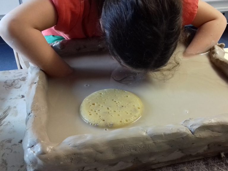 Hände in Wasserschüssel Ein Kind beugt sich über ein Tonfeld, das halbvoll mit Wasser gefüllt ist. Die Hände sind eingetaucht, wir sehen nur die Haare des Kindes.
